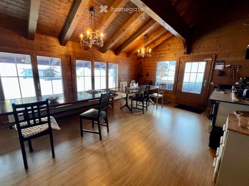 Cozy log cabin interior with exposed wooden beams, large windows offering a scenic view, dining area with a table and chairs, and a small kitchen area with a counter and appliances.
