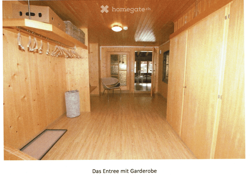 A narrow hallway with wooden walls and flooring, featuring built-in coat racks, a small chair, and a trash bin. The hallway leads to an open doorway with glass panels.