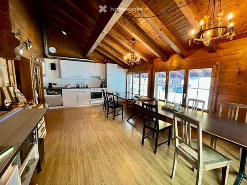 Bright, open-plan living and dining area with exposed wooden beams, white kitchen cabinets, a dining table with chairs, and a chandelier hanging from the ceiling.