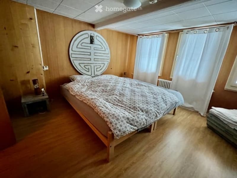 Cozy bedroom with wooden walls and floor, a double bed with patterned bedding, a nightstand with a lamp, and a radiator near the window with white curtains.