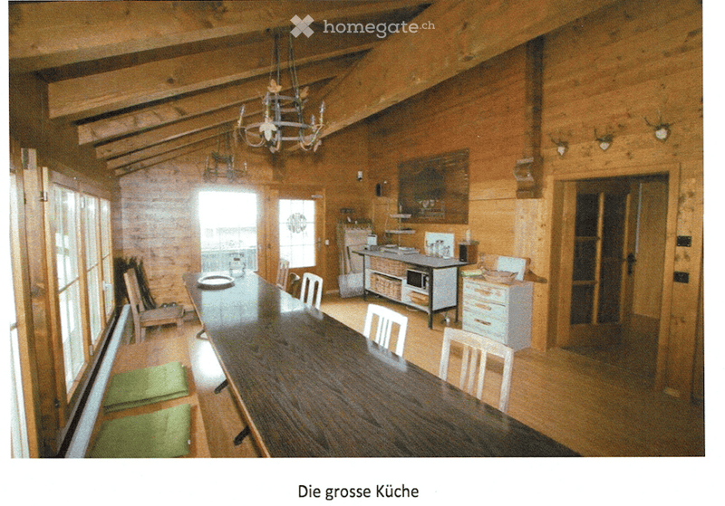 A spacious, wooden-paneled dining area with a long wooden dining table surrounded by chairs, a small kitchen area with cabinets and a microwave, and a chandelier hanging from the ceiling.