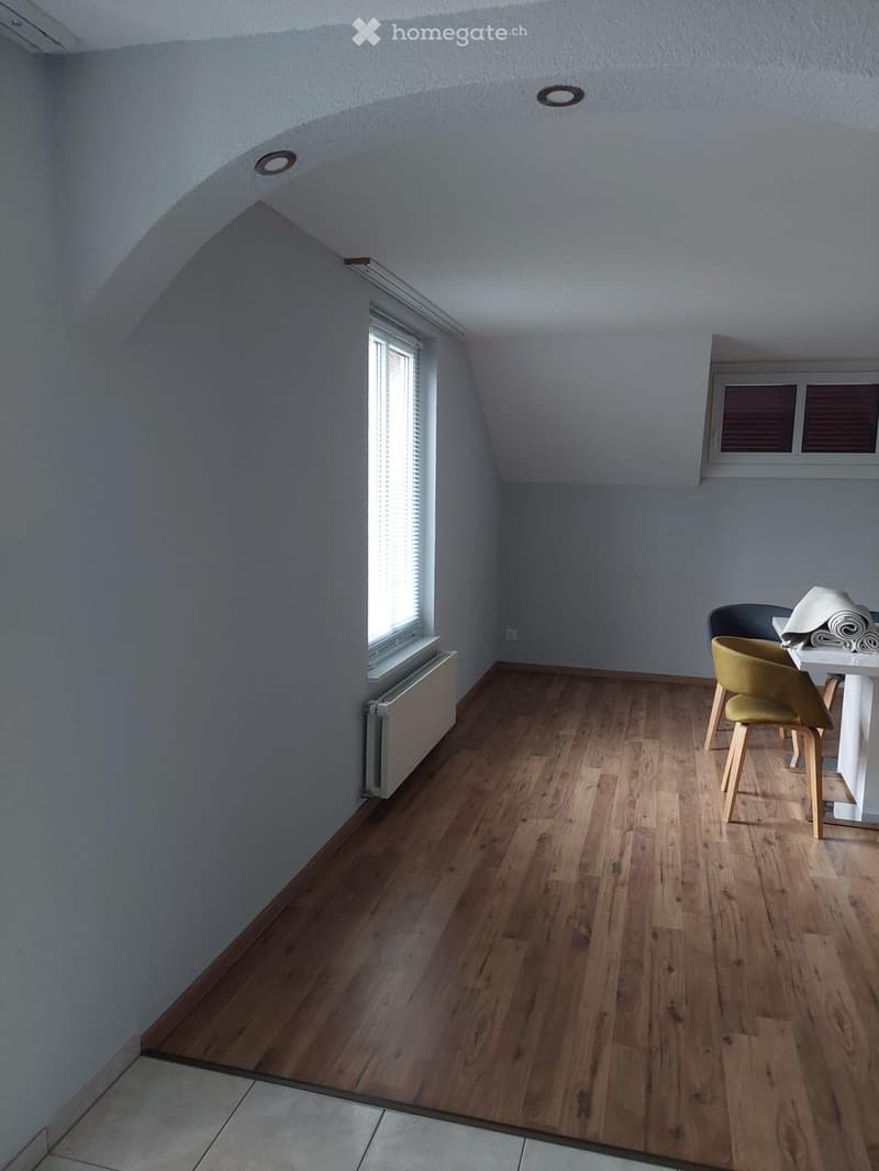 Bright attic room with wooden flooring, slanted ceiling, and two windows. Contains a small table and two chairs.