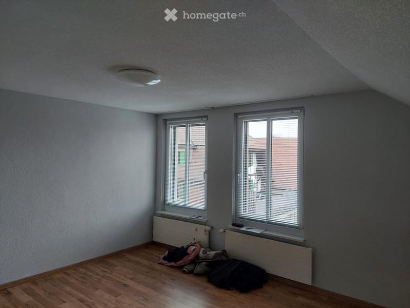 Empty room with wooden flooring, two windows with blinds, radiator, and some scattered items on the floor.