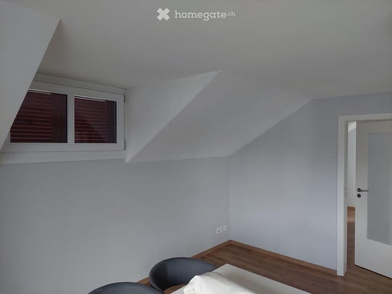 A small, unfurnished room with light blue walls, wooden flooring, and a slanted ceiling. A single window with blinds is on one wall, and a door leads to another room.