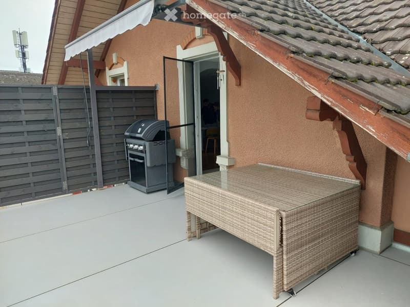 Outdoor patio with a wicker table and a grill, adjacent to a house with a covered entrance.