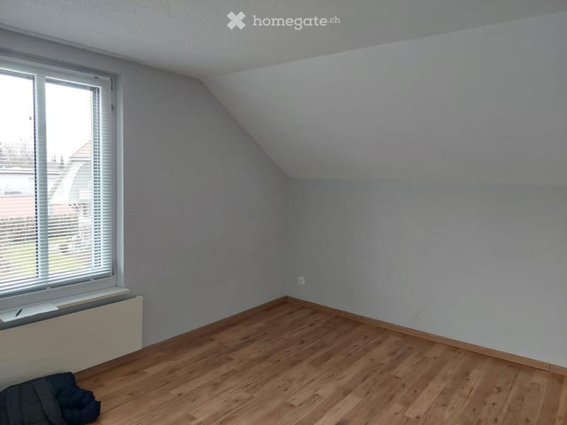 Empty attic room with wooden flooring, slanted ceiling, and a single window with blinds. There is a radiator below the window and a small pile of items on the floor.