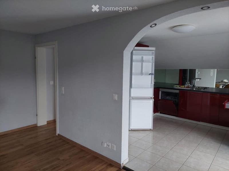 A small, modern kitchen area with a refrigerator, red cabinets, a sink, and a faucet. The space features wooden flooring on one side and tiled flooring on the other, with a white pillar separating the areas.