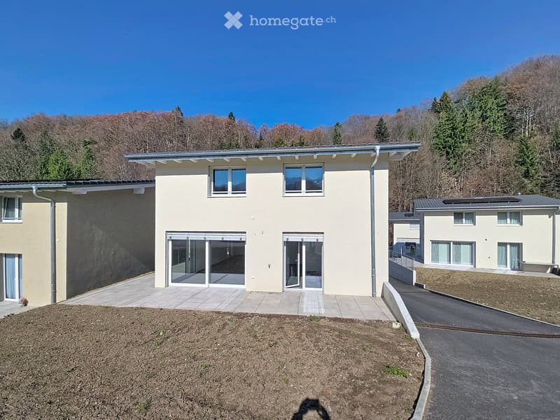 Modern two-story house with light yellow facade, large windows, and sliding glass doors leading to a paved patio area. The house is situated on a raised ground floor with a driveway and surrounded by trees and hills.