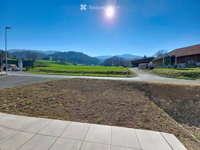 A landscaped outdoor area with a paved walkway, grassy slope, and distant rural buildings and mountains under a clear blue sky.