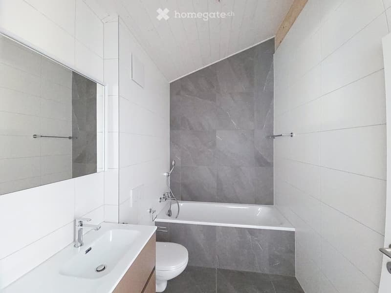 Modern bathroom with white tiles, a large mirror, a sink, a toilet, and a bathtub with a grey accent wall.