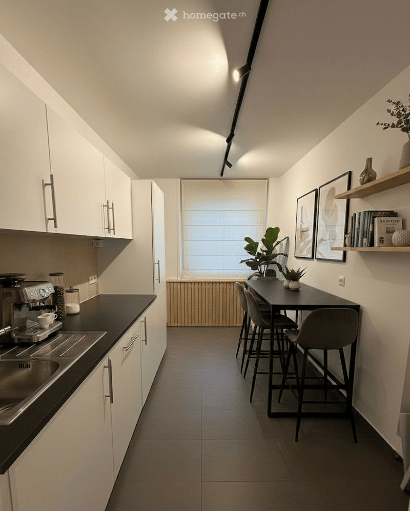 Modern kitchen with white cabinets, black countertops, and a small dining area. Features include a sink, coffee maker, bar stools, a small table, and decorative plants.