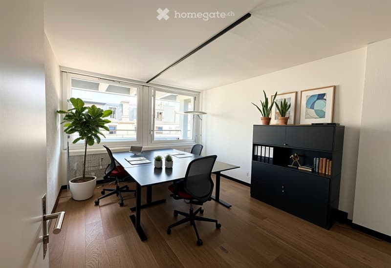 Modern office space with a large wooden desk, ergonomic chairs, a dark cabinet, potted plants, and framed artwork on the wall. Large windows provide natural light.