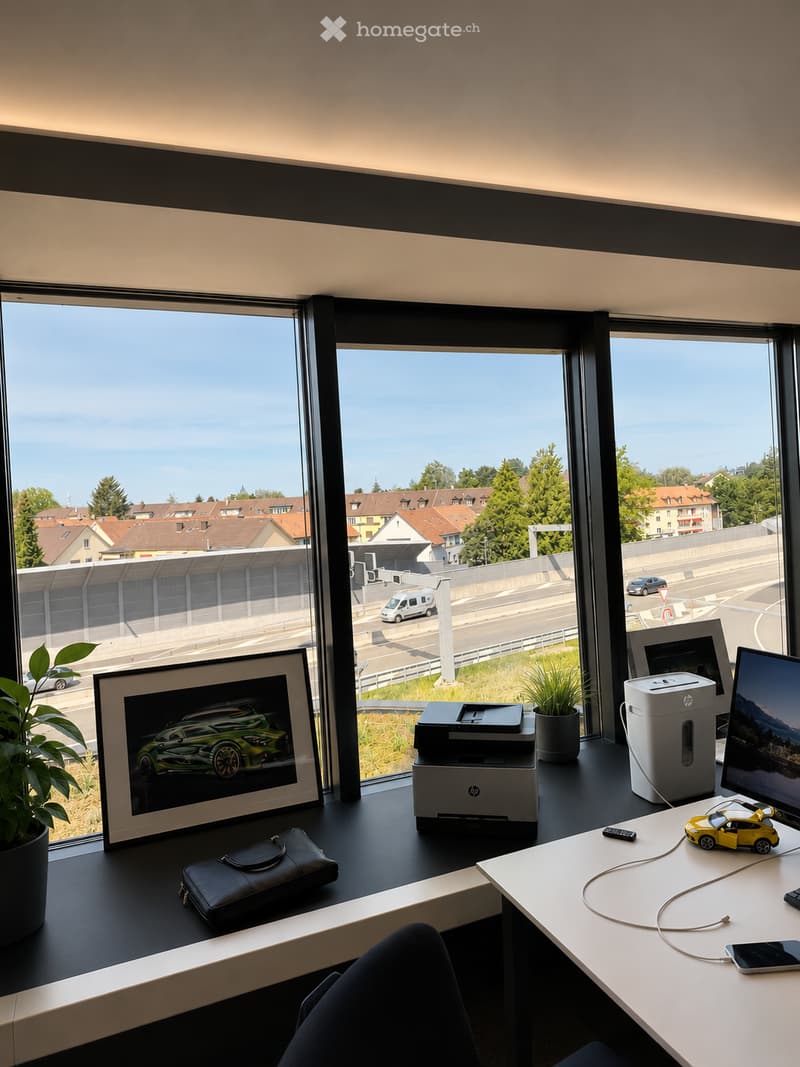 Modern office space with large windows overlooking a cityscape, featuring a desk with a printer, screen, laptop, cellphone, picture frame, plant, and a small toy car.