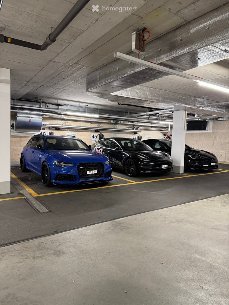 Underground parking area with several parked cars, including a blue Audi and a black Tesla, marked parking spaces, and visible structural elements like pillars and lighting.
