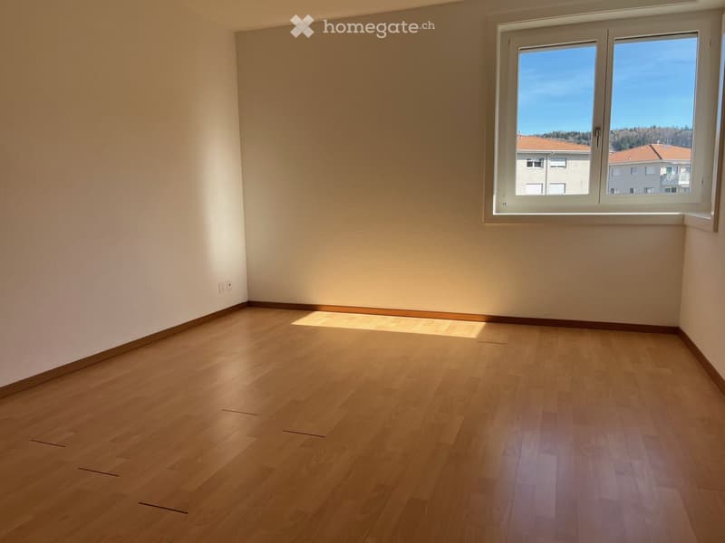 Empty room with light wooden flooring and white walls, featuring a large window with a view of neighboring buildings and a clear blue sky.