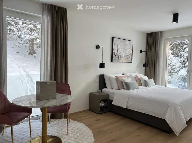 bedroom with a bed, nightstand, two chairs, table, rug, and large windows with snowy view