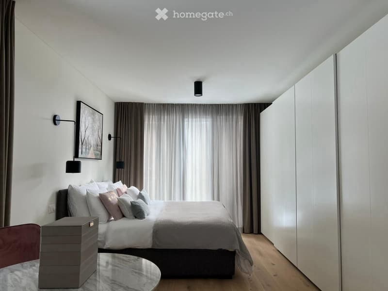 Bedroom with bed, white walls, wooden floor, large windows, wall-mounted lamps, picture frame, white and grey bedding, white wardrobes, round table with storage box
