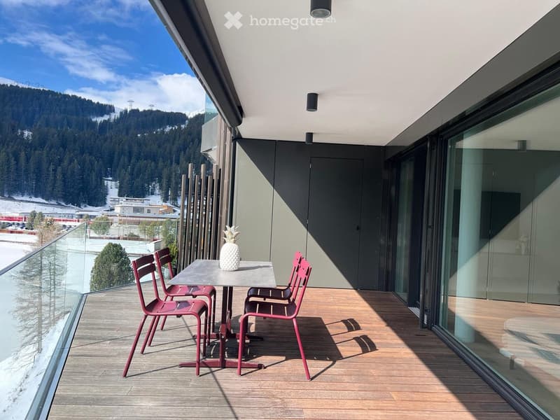 Balcony with wooden floor, glass railing, table with chairs, and a view of the snowy mountains