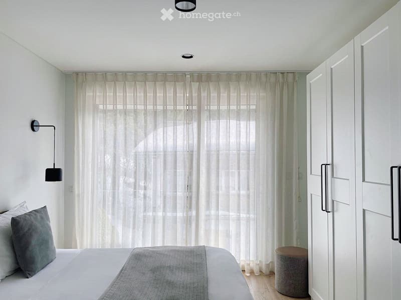 Bedroom with white walls, wooden floor, bed, white curtains, wall lamp, white and gray pillows, white wardrobe, ottoman