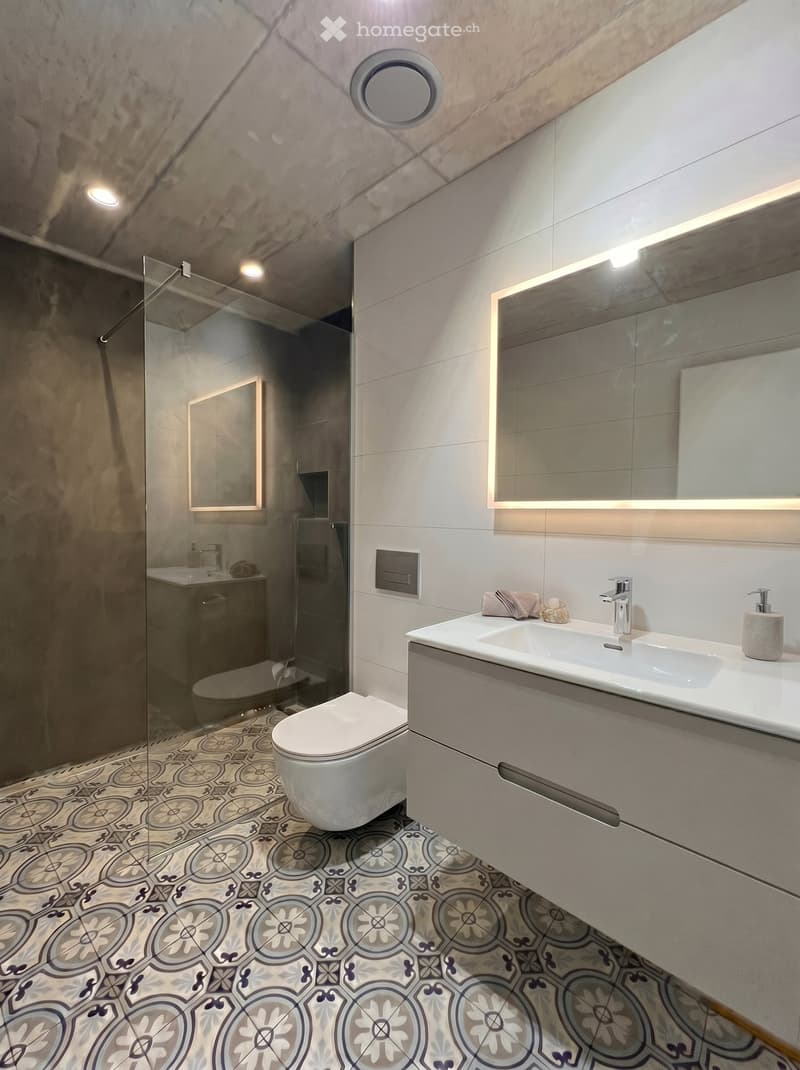 Modern bathroom with a white sink, chrome faucet, large mirror with built-in lighting, toilet, glass-enclosed shower, and decorative patterned tiles.