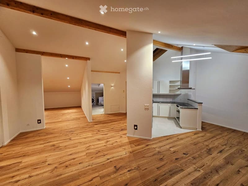 An unfurnished room with wooden flooring, white walls, and exposed wooden beams on the ceiling. The room features a small kitchen area with white cabinets and a countertop, and a separate laundry area with a washing machine and dryer visible in the backgr