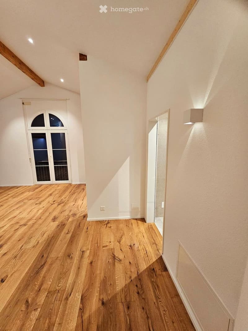Empty hallway with wooden flooring, white walls, and recessed lighting. Features a large arched window and a partially visible door frame on the right side.