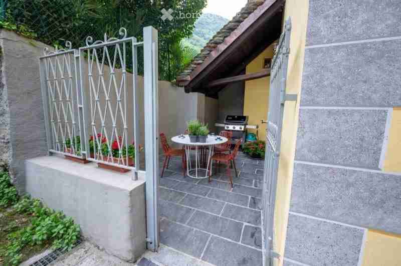 Outdoor patio with a round table, four chairs, a potted plant, and a grill. The patio is enclosed by a metal gate and has a tiled floor.