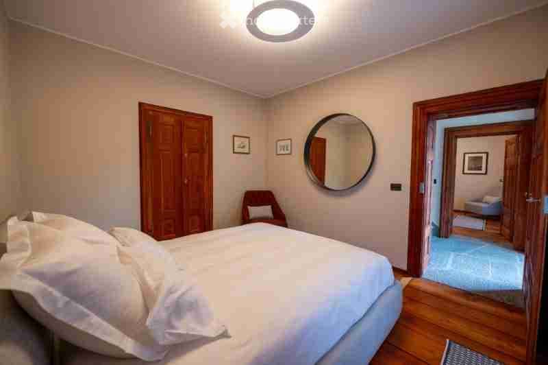 Cozy bedroom with a large bed, wooden wardrobe, a small chair, and a round mirror on the wall. The room features light-colored walls and wooden flooring.