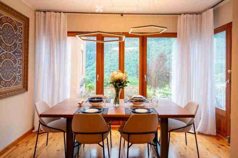 Bright dining room with large windows offering a scenic view, a wooden dining table set with plates, glasses, and a vase of flowers, surrounded by modern chairs.