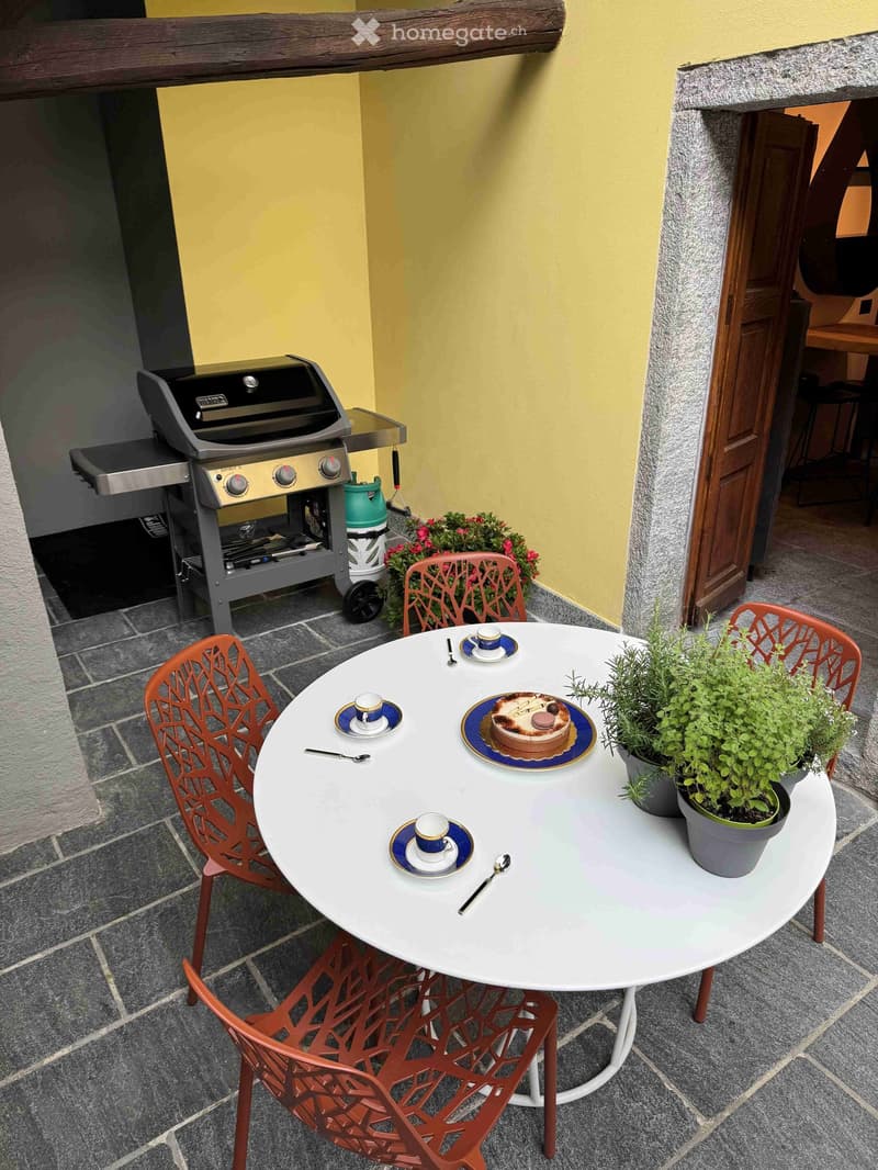 Balcony with a round white table set for four, red metal chairs, potted plants, and a grill in the background.