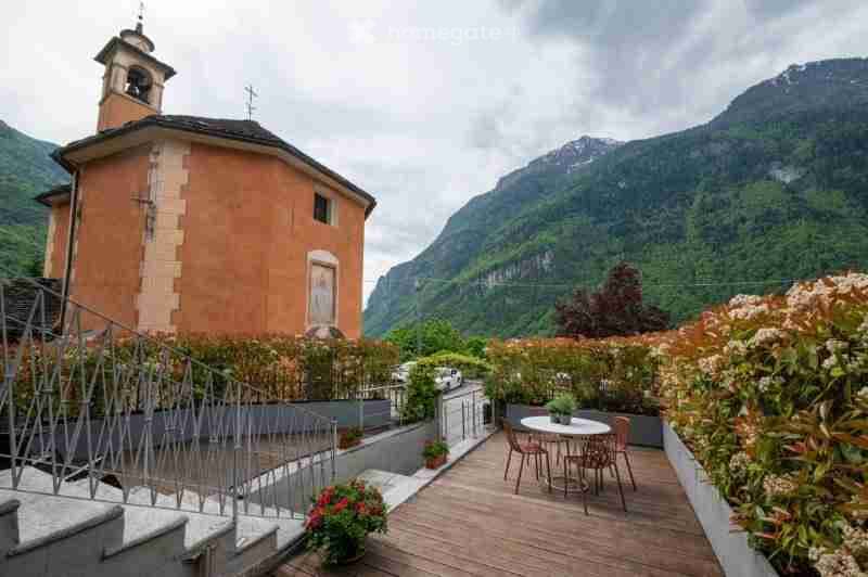 A wooden terrace with a small table and chairs, surrounded by potted plants and flowers, overlooking a scenic mountain view and a small chapel.