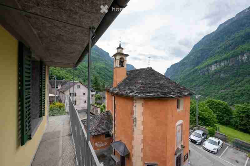 Balcony overlooking a picturesque village with a church tower, surrounded by mountains and greenery.