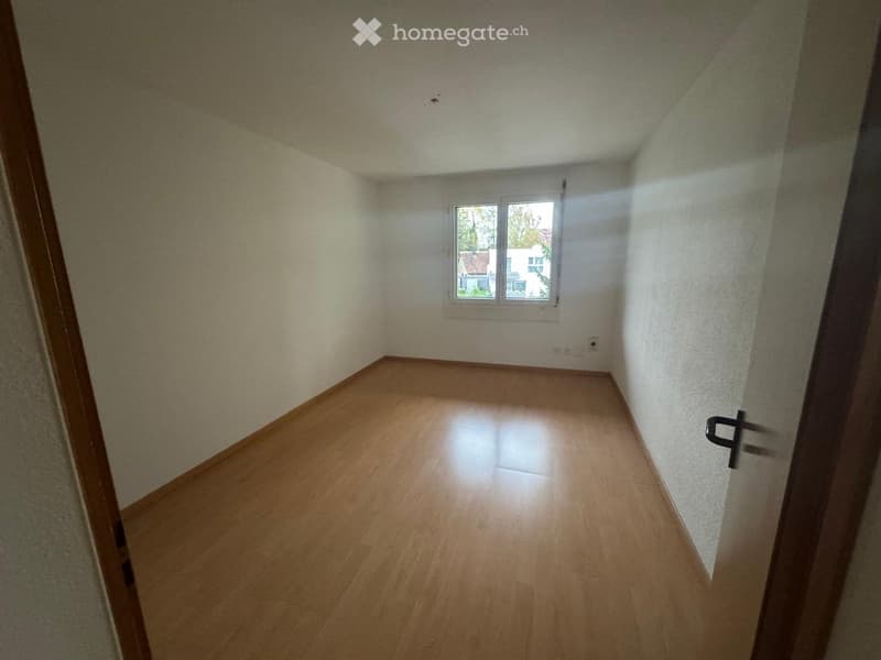 Empty room with wooden flooring, white walls, and a single window showing a view of neighboring buildings.