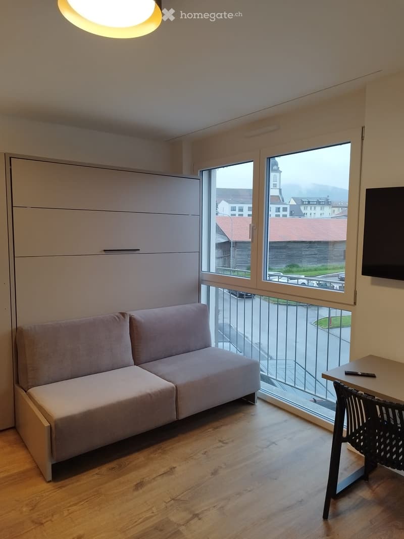 Modern living room with a grey sofa, a small table with a chair, and a TV mounted on the wall. Large windows offer a view of the city and a balcony area.