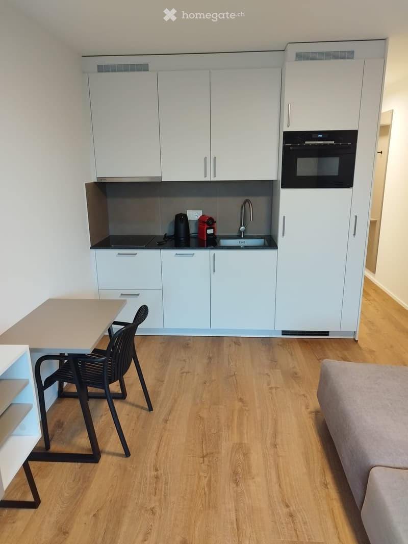 Modern, open-plan living and dining area with a kitchen. Features include light-colored wooden flooring, white kitchen cabinets, a built-in oven, a sink with a modern faucet, a coffee maker, a dining table with a black chair, and a light-colored sofa.