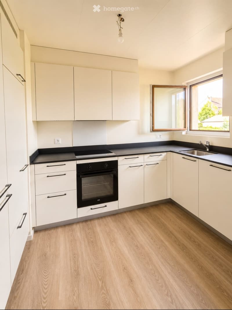 Modern kitchen with white cabinets, black countertops, and a built-in oven. Features a wooden floor and large windows letting in natural light.