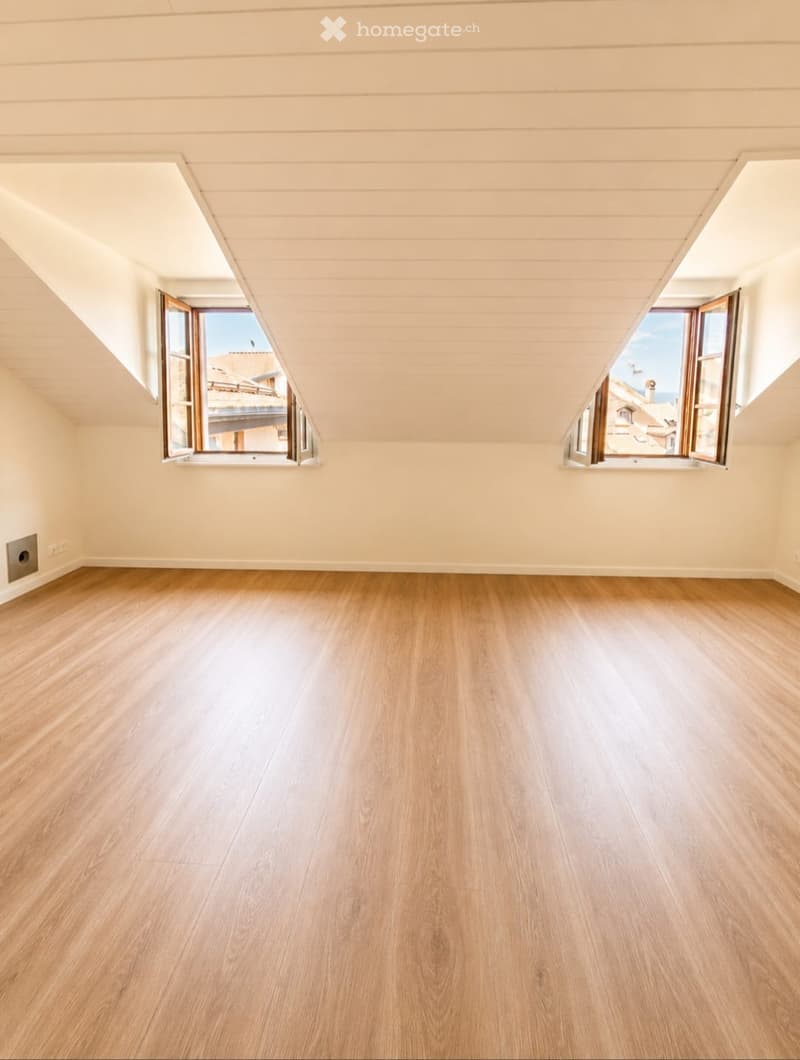 Empty attic room with wooden flooring, slanted ceilings, and two large windows offering a view of the outside.