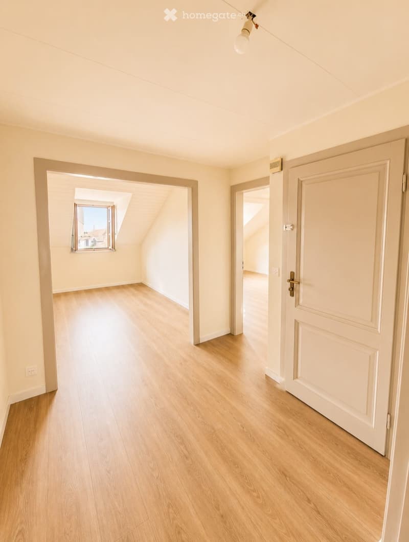 Empty room with light wooden flooring, white walls, and a closed white door. Natural light streams in from a window in an adjacent room.