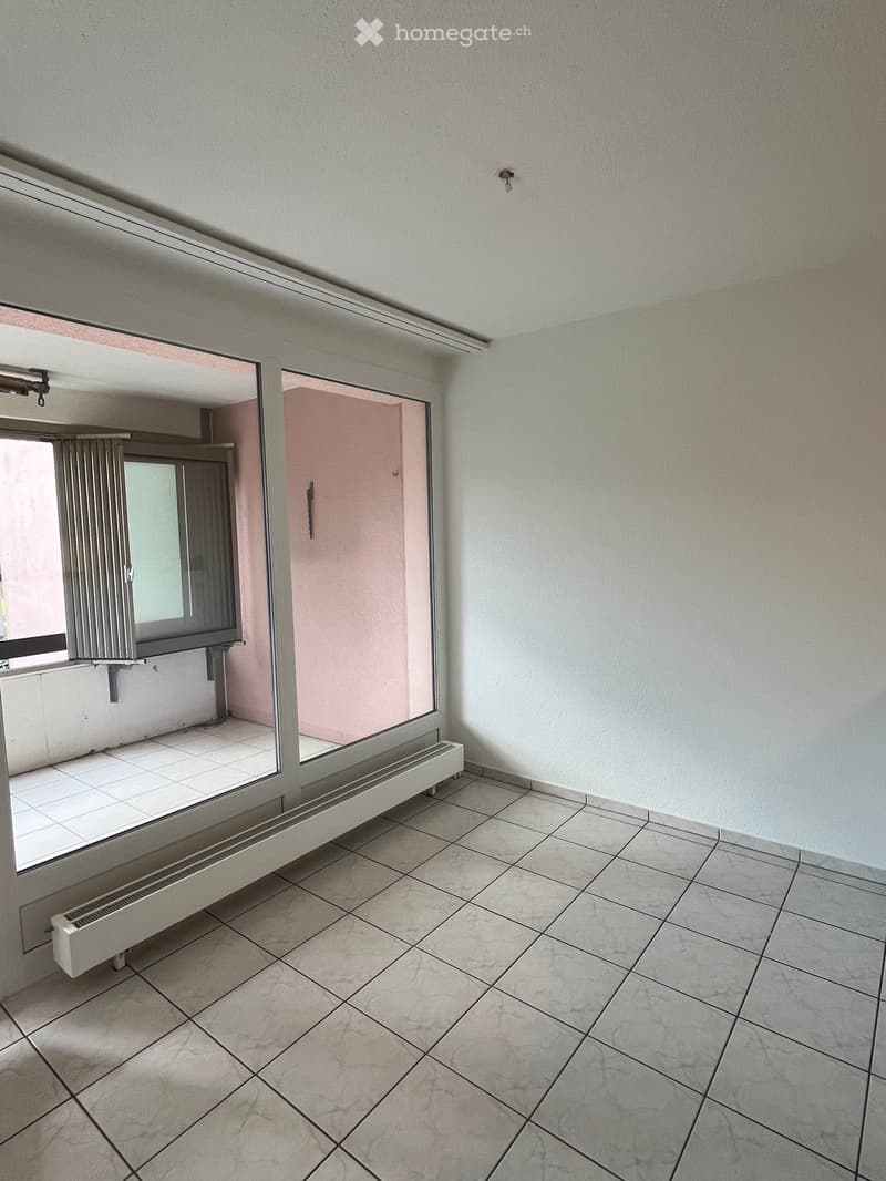 Empty room with tiled floor, white walls, and a large window with radiator below. Adjacent room visible through doorway.