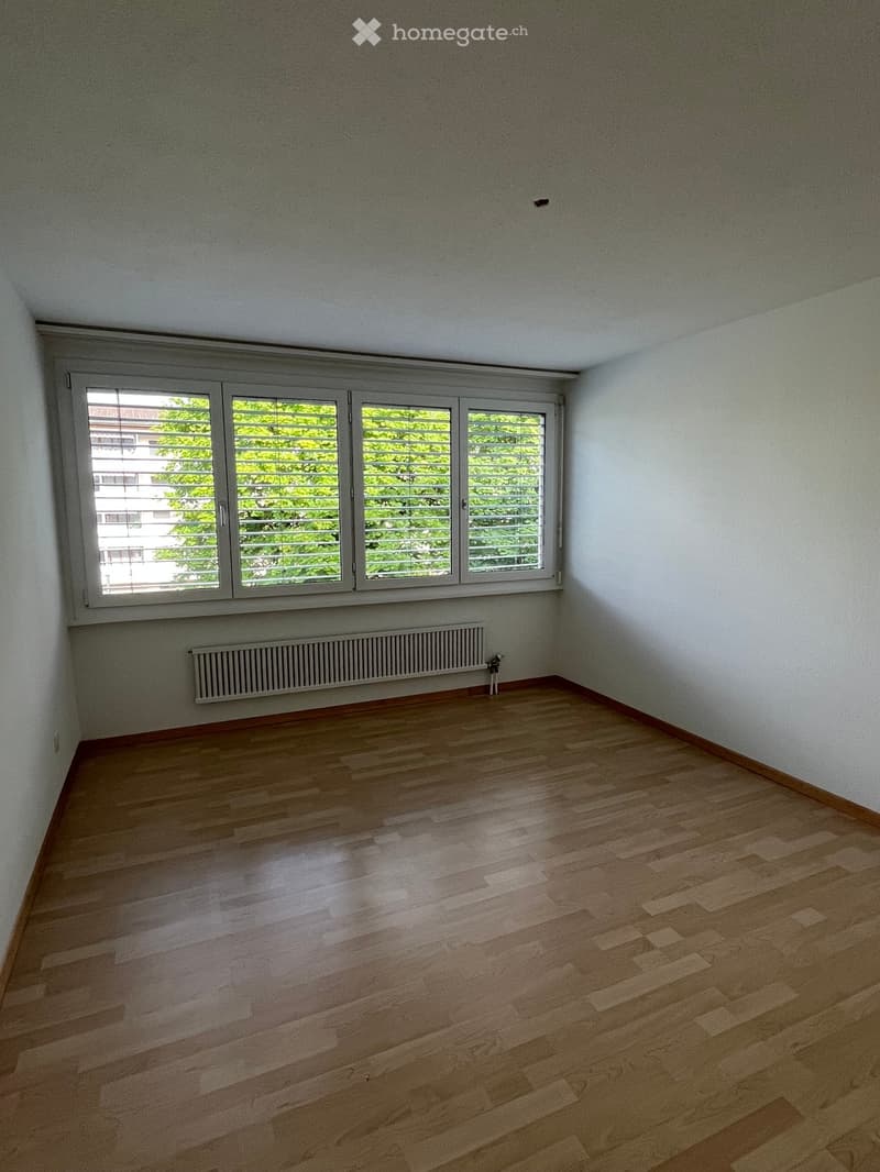 Empty room with wooden flooring, white walls, large window with blinds, and radiator below the window.