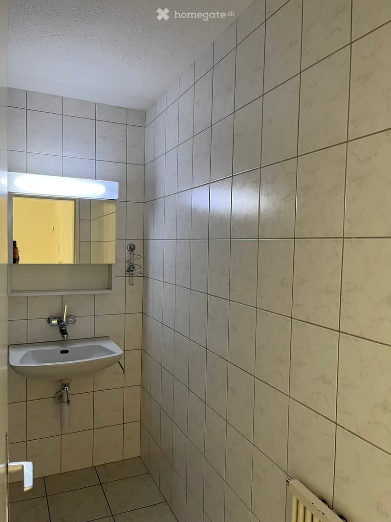 Small bathroom with beige tiled walls and floor, a white sink with a chrome faucet, a mirror with built-in lighting, and a radiator.