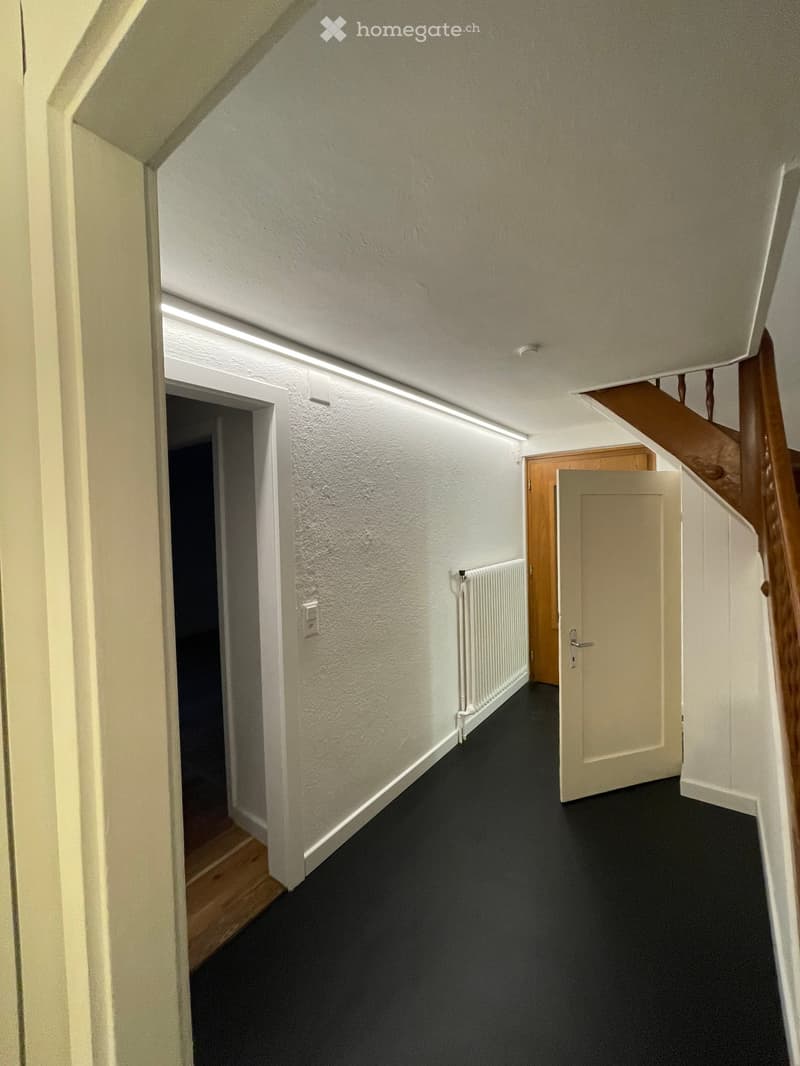 A narrow hallway with white walls, black carpeted floor, and a wooden staircase leading upwards. There are doors on both sides and a radiator on one wall.