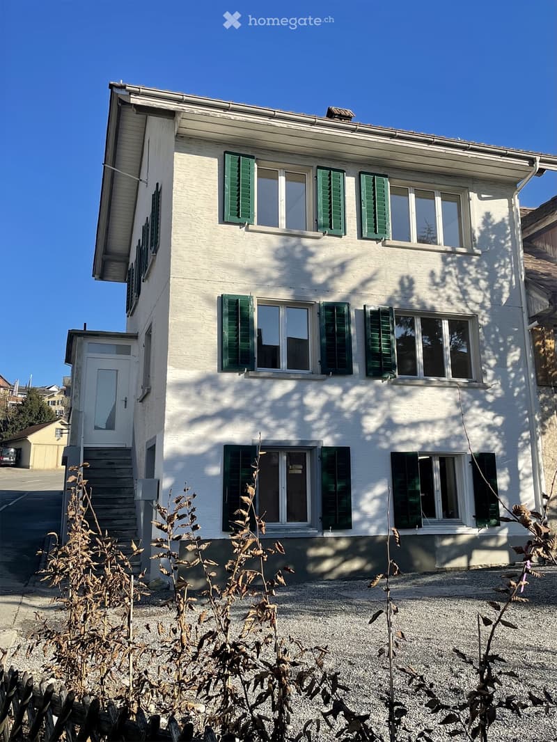 Two-story white house with green shutters, multiple windows, and a small entrance door. The house is surrounded by gravel and some dry plants.