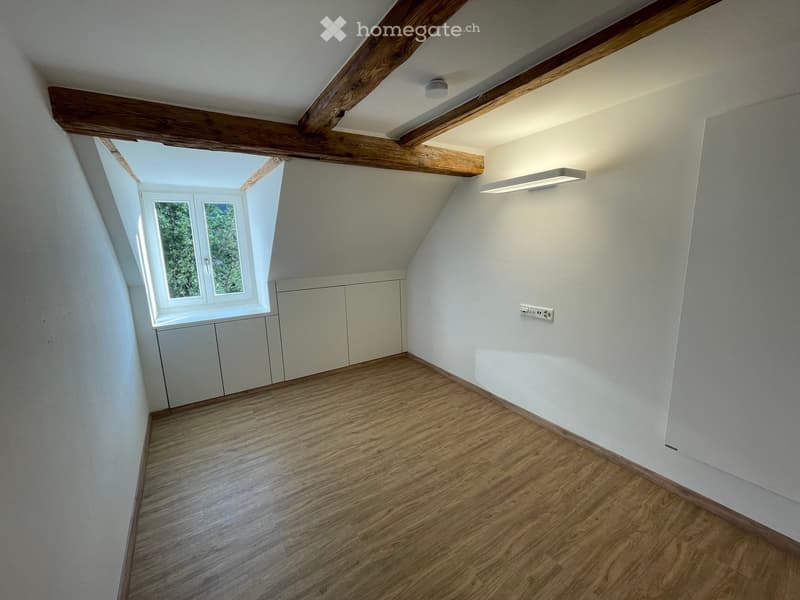 Empty attic room with wooden floor, white walls, exposed wooden beams, and a skylight window. Minimal built-in cabinetry is visible.