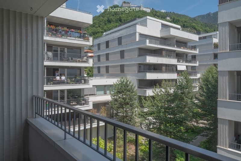 Modern multi-story residential complex with balconies, overlooking a lush green hillside and distant mountains.