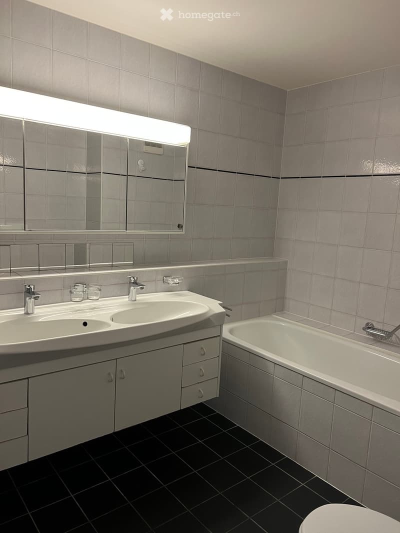 Modern bathroom with grey tiled walls, black tiled floor, double sink with chrome faucets, large mirror with built-in lighting, bathtub, and toilet.