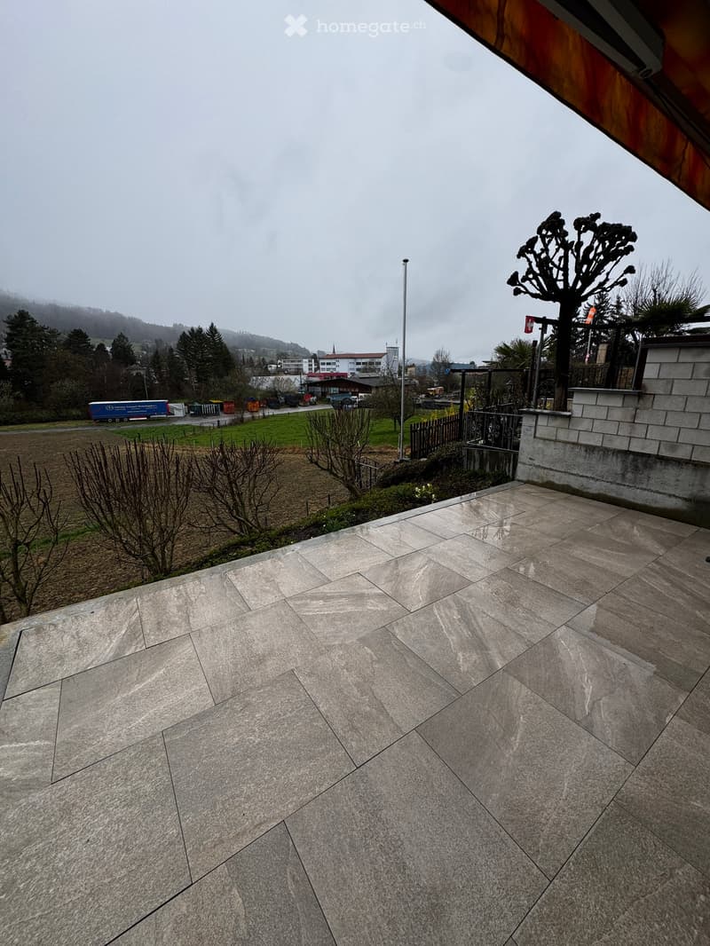 A spacious outdoor patio with large, light-colored stone tiles, surrounded by a low stone wall, trees, and a view of a rural landscape with fields and distant hills.