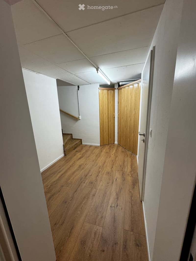 Empty hallway with wooden flooring, white walls, and a staircase leading upwards. Wooden doors are visible at the end of the hallway.