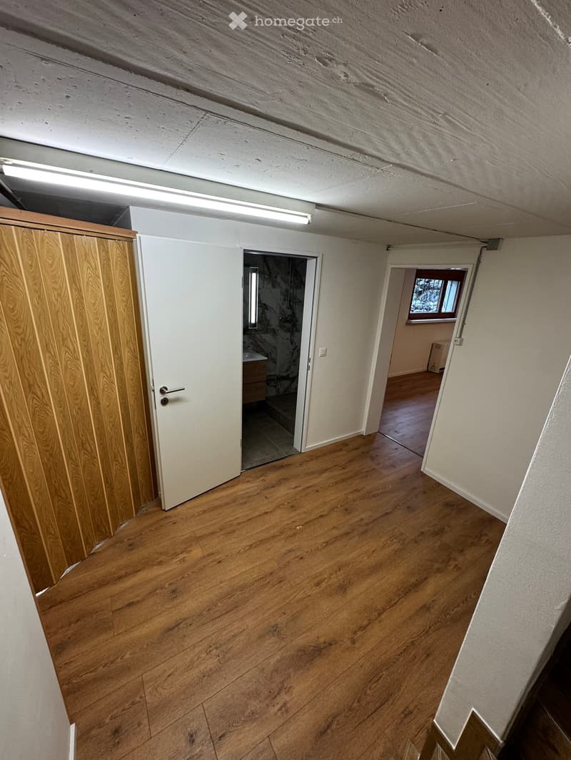 Empty room with wooden flooring, white walls, and a wooden door. A small window is visible in the adjacent room.
