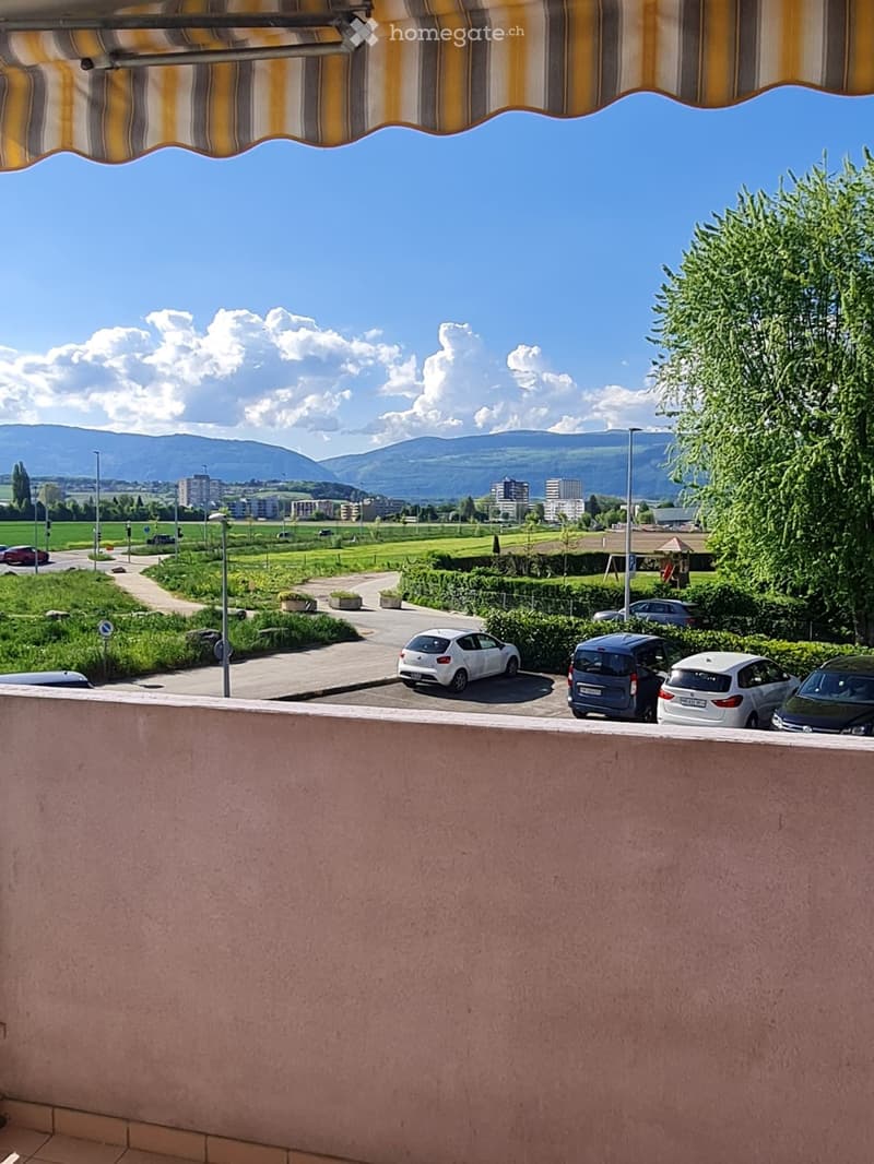 Outdoor balcony with a scenic view of the mountains, parking lot, cars parked, lush greenery, trees, and a clear blue sky